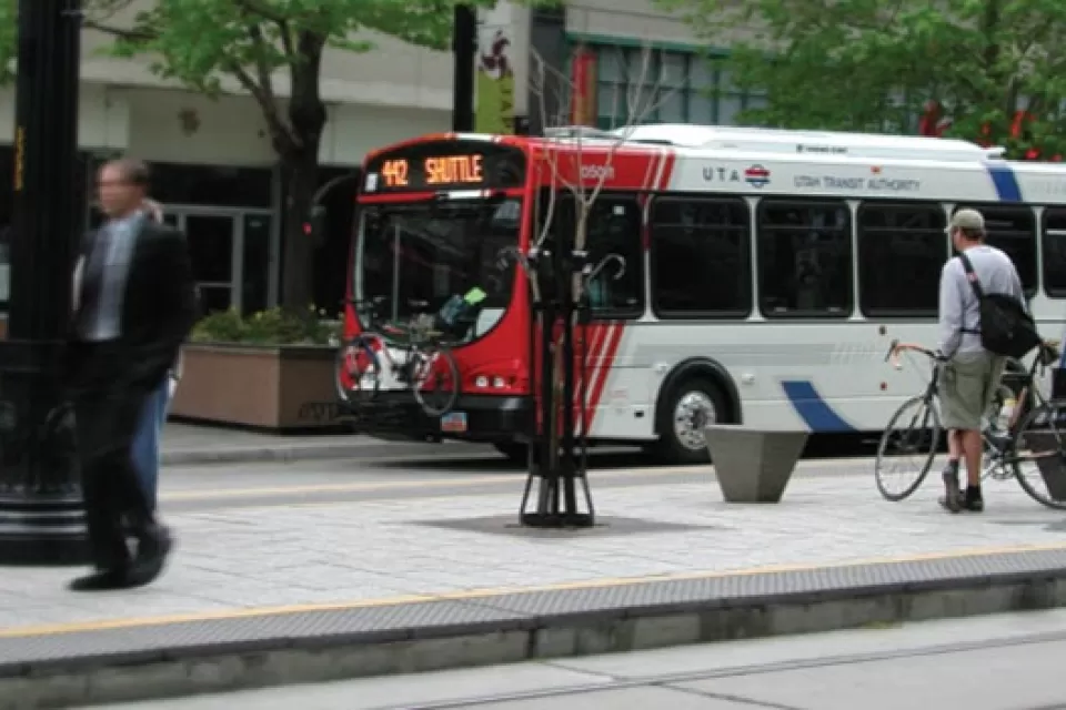 White, red, and blue Utah Transit Authority bus at stop with man standing on sidewalk next to bicycle